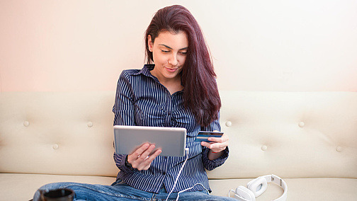 Junge Frau sitzt mit dem Tablet auf der Couch und vergleicht ihre Kreditkarteninformationen.
