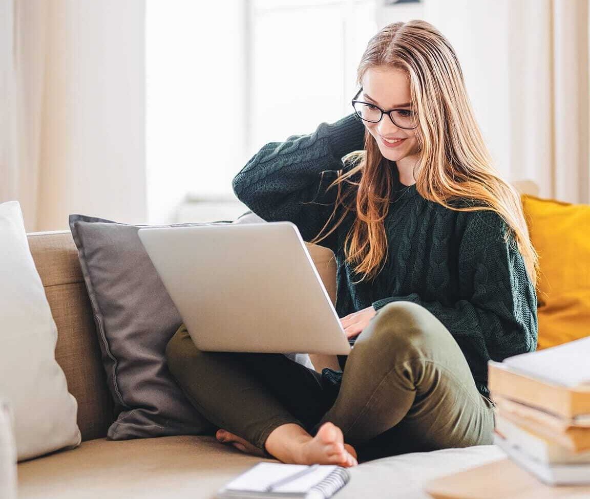 Jugendliche sitzt im Schneidersitz auf der Couch und hat einen Laptop im Schoß.