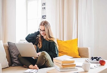 Jugendliche sitzt im Schneidersitz auf der Couch und hat einen Laptop im Schoß.