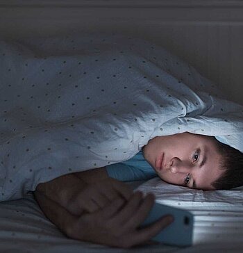 Junge unter Bettdecke schaut müde auf Smartphone