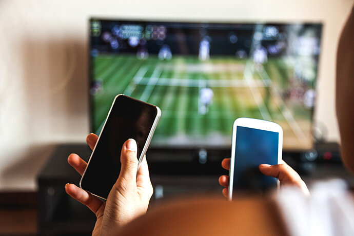 Zwei Jugendliche mit Smartphones in der Hand. Im Hintergrund läuft ein Fußballspiel im TV.