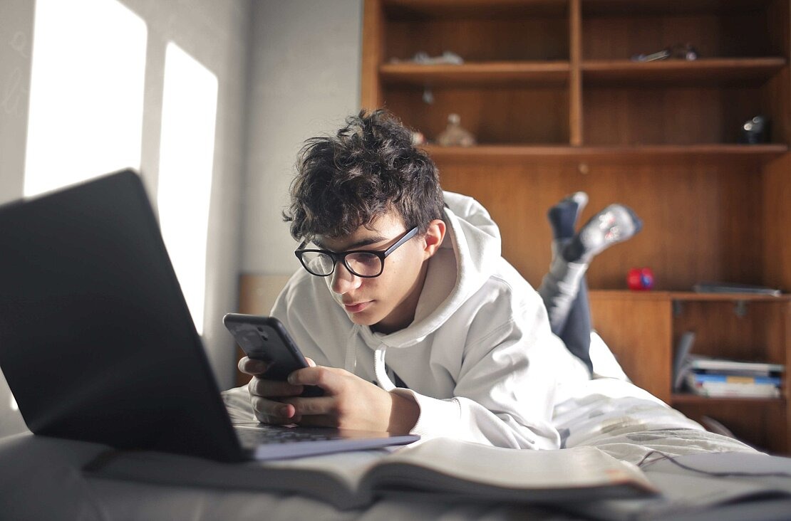 Junge schaut auf Handy und hat gleichzeitig den Laptop geöffnet