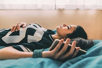 Teenagerin liegt mit Smartphone auf dem Bett und guckt betrübt an die Decke.