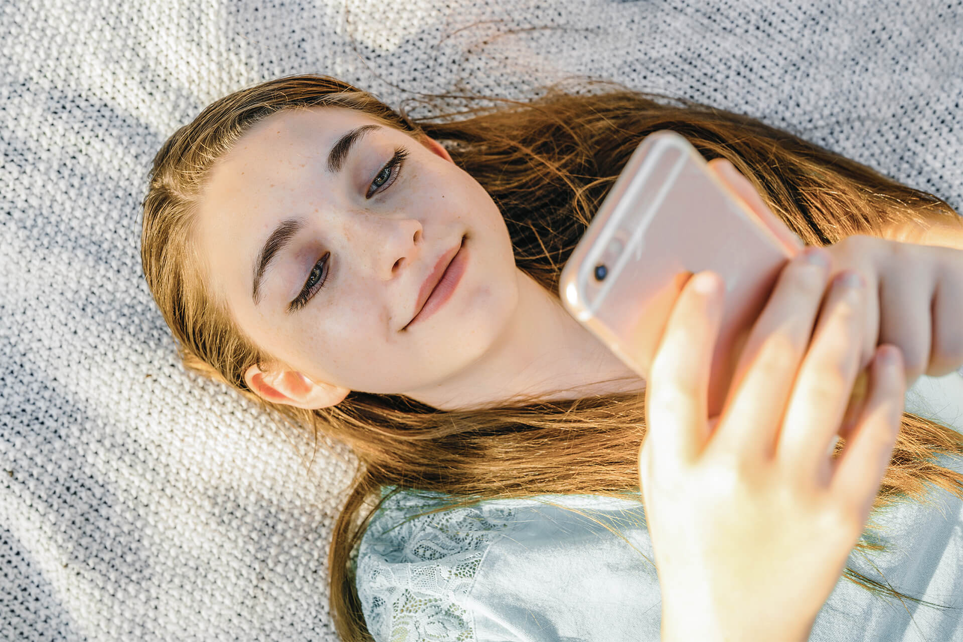 Mädchen liegt mit dem Rücken auf einer Decke und schaut hoch zu ihrem Handy.